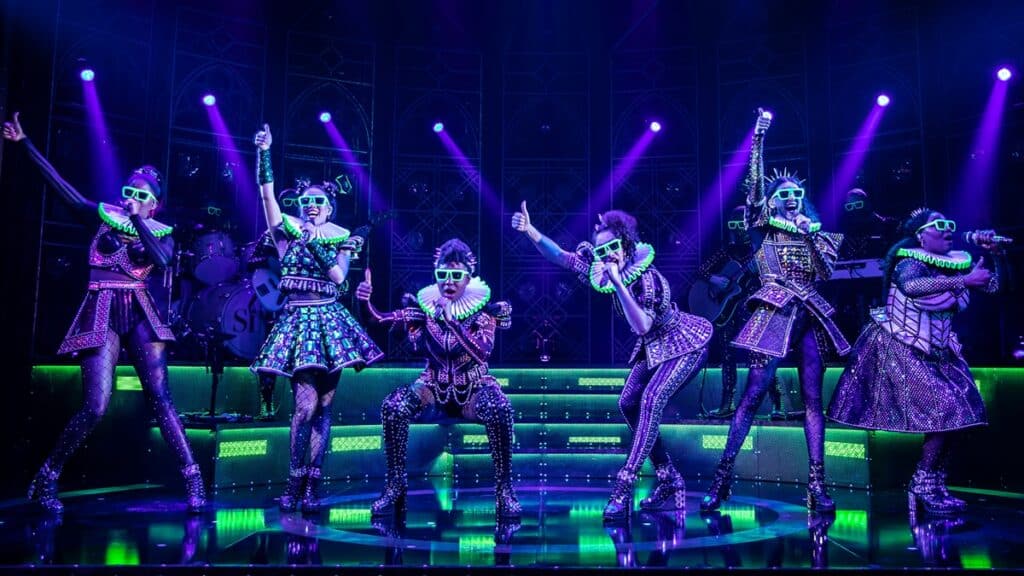 The six queens from Six the Musical sing into microphones wearing neon green glasses.