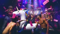 A woman rides an inflatable unicorn, surrounded by male strippers and a rain of pink dollar bills for Magic Mike Live