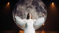 Paris Paloma raises her hands in front of front of a giant moon