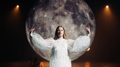 Paris Paloma raises her hands in front of front of a giant moon