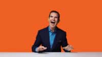 Jimmy Carr laughs, sitting at a table