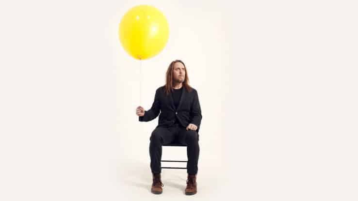 Tim Minchin sits holding the string of a large balloon