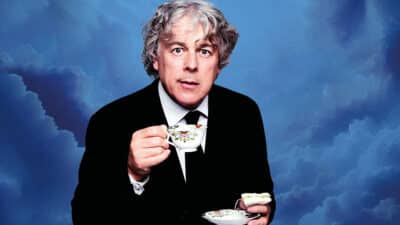 Alan Davies holds a teacup, wearing a dark suit