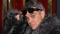 Robbie Williams lifts his sunglasses, wearing a fur coat.