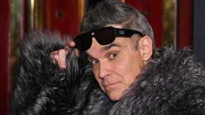 Robbie Williams lifts his sunglasses, wearing a fur coat.