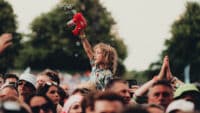 A child at a festival sits on someone's shoulders as he wantonly fires a bubble gun into the air