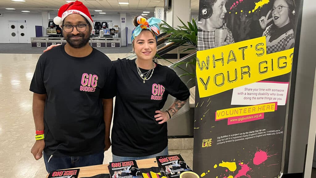 A man in a santa hat stands next to a woman at a gig venue, handing out leaflets to promote Gig Buddies from Stay Up Late