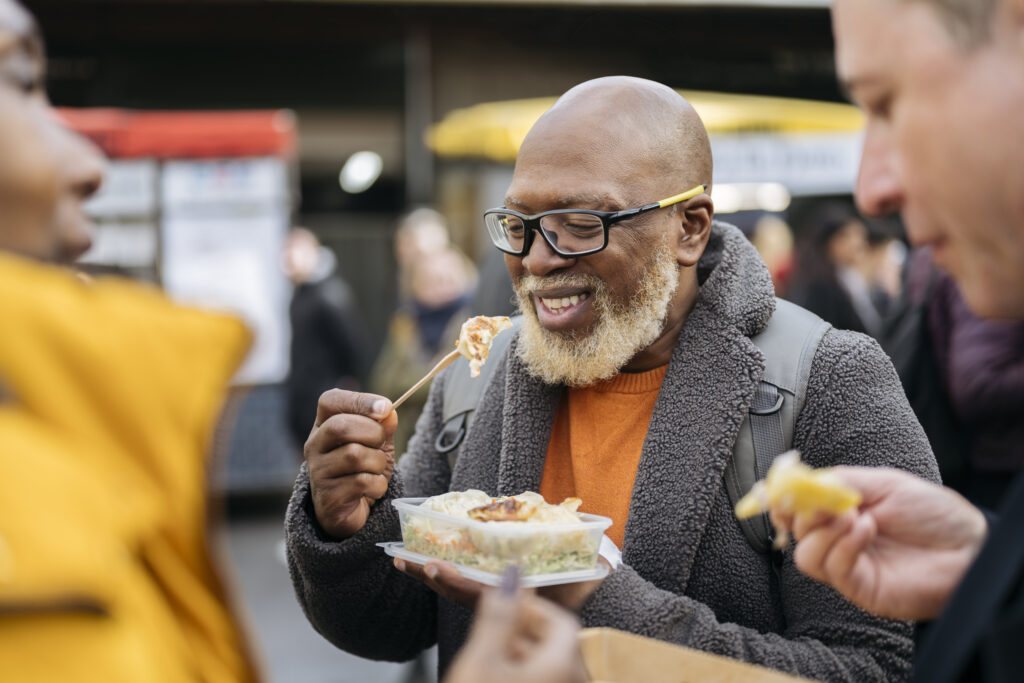 Mature Black couple and Caucasian friend talking and eating as they spend weekend leisure together at Southwark market hall.