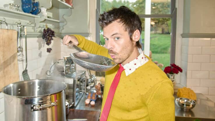 Harry Styles stands over a cooking stove, blowing on a giant spoon to cool its contents.