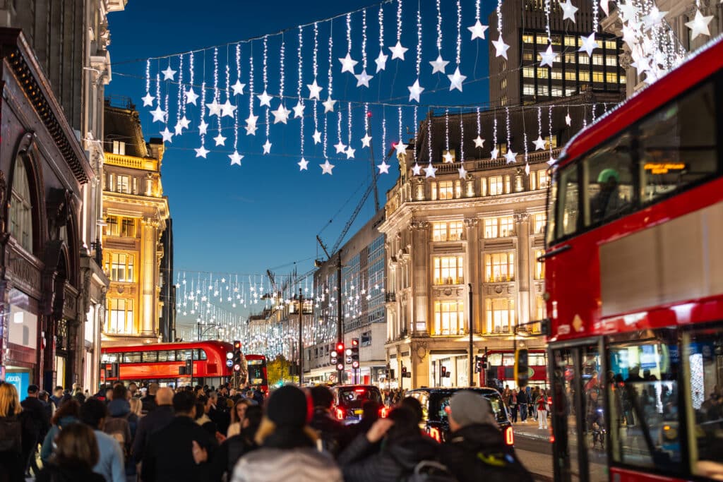 Christmas star lights illuminating a busy Oxford Street with shoppers, red buses, and city buildings during the evening
