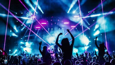 Rear view of excited crowd enjoying a DJ performance at a festival. There are many raised hands, some of the holding cell phones and taping the show